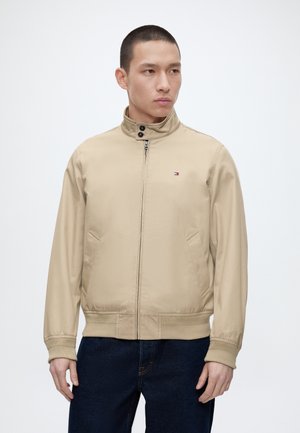 Mand iført beige lynlåstrøje med stående krave og mørke jeans, kigger til siden mod en ensfarvet hvid baggrund.