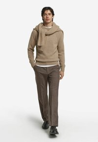 Beige stickad tröja med ribbat mönster, bärs över en vit skjorta. Matchas med bruna rutiga byxor. Svarta skor fulländar outfiten.