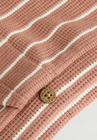 Texturerad terrakotta tyg med vita ränder, med en rund brun knapp märkt "NEXT." Nära bild av stickmönstret.