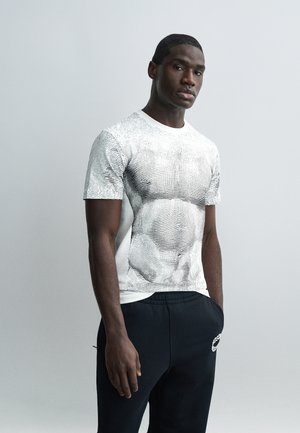 Fiatal férfi áll egyszínű háttér előtt, fehér pólót visel fekete absztrakt vonalakkal és fekete nadrágot, kezét zsebében.