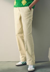 Crème kleurige broeken met rechte pijpen, zijzakken en subtiele textuur, gecombineerd met sportieve groen-witte sneakers.