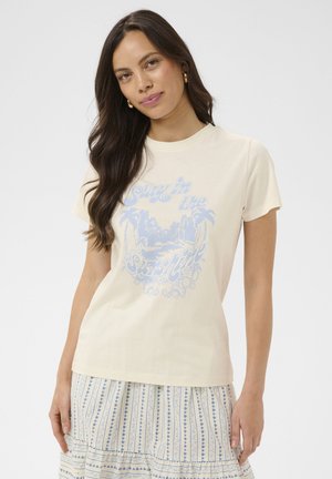 Ženska z dolgimi temnimi lasmi nosi kremno majico s svetlomodrim napisom "Surf in the Sunshine" in vzorcem palme, skupaj z vzorčasto krilo.