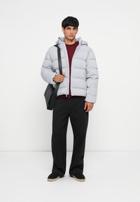 Light gray puffer jacket with a zip front, elastic cuffs, and a hood, worn over a maroon shirt, with black trousers and a black bag.