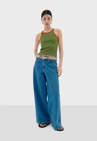 Zelený pletený top bez rukávů s oranžovým lemováním, kombinovaný s kalhotami širokého střihu z modrého denimu a vzorovaným pásem, stylizovaný s černými žabkami.
