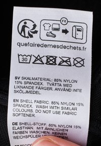 Vaatteiden hoito-ohjeet valkoisella kankaalla, sisältäen hoito-ohjeita useilla kielillä, sekä symboleita, jotka osoittavat pesu-, kuivaus- ja hävittämismenetelmiä.