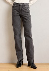 Jean en denim gris avec une coupe droite, cinq poches et une fermeture à bouton, associés à des bottes à talons noirs brillantes.