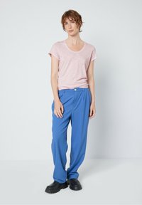 Gaiši rozā īsās piedurknes t-krekls, kas savienots ar brīviem, spilgti zilajiem biksēm un melniem klogiem, uz vienkārša baltā fona.