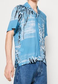 Koszula z krótkim rękawem wykonana z lekkiej niebieskiej tkaniny, ozdobiona abstrakcyjnymi białymi i czarnymi wzorami, z kołnierzykiem typu camp i luźnym krojem.