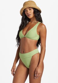 Bikini verde claro con textura acanalada, que presenta una parte superior triangular y un fondo de cintura alta. La modelo lleva un sombrero de paja y collares en capas.