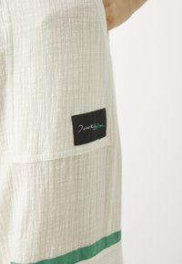 Hvit teksturert stoff med en grønn vannrett stripe og en svart etikett med grønn tekst. Designet inkluderer en lomme.