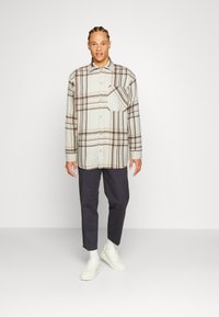 Camicia a quadri color beige chiaro con motivo a tartan marrone, chiusura con bottoni, taschino sul petto, abbinata a pantaloni scuri e sneaker bianche.
