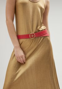 Guldmetallisk klänning med ett flödande material, åtdragen med ett brett röd läderbälte som har en cirkulär guldspänne med ett hjärtformigt mönster.