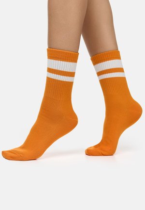 Benen met heldere oranje korte sokken met twee witte horizontale strepen bij de bovenkant, afgebeeld tegen een effen witte achtergrond.