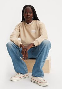 Sudadera beige con el logo "Levi's" en blanco, combinada con jeans baggy de color azul claro y zapatillas beige con acentos en rojo y azul marino. Textura suave del tejido.