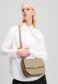 Borsa a tracolla khaki realizzata in materiale morbido con chiusura in tonalità oro. Presenta bordi curvi e una tracolla per il trasporto.