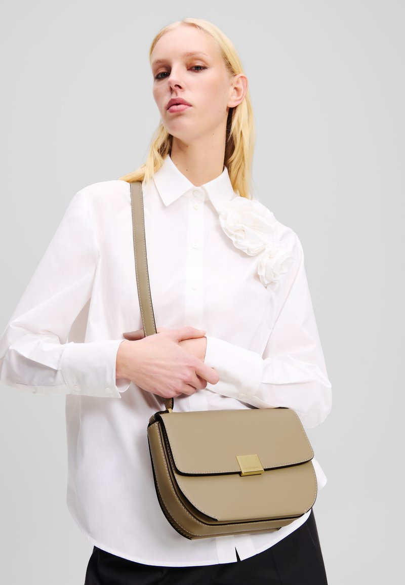 Borsa a tracolla khaki realizzata in materiale morbido con chiusura in tonalità oro. Presenta bordi curvi e una tracolla per il trasporto.