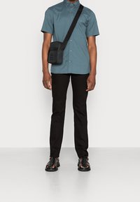 Maglietta a maniche corte teal con colletto button-down, pantaloni neri slim-fit, borsa a tracolla nera e sandali neri con aperture.
