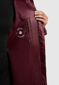 Burgundi puffertakó fényes steppelt textúrával és logó részlettel. A belső részen oldalsó zseb és elegáns befejezés található.