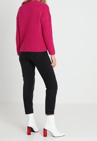 Donna che indossa un pullover rosa brillante, jeans neri e stivaletti bianchi con tacco rosso, in piedi davanti a uno sfondo bianco uniforme.
