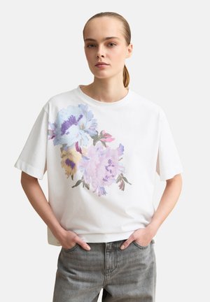 Joven con piel clara y cabello recogido, vistiendo una camiseta blanca con un gran estampado floral en tonos pastel y jeans grises, con las manos en los bolsillos.