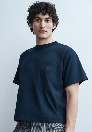 Nooruk meest käänuliste juustega, kes kannab tumesinist Versace rinnataskuga T-särki ja triibulisi pükse, seistes ühevärvilise heleda taustaga.