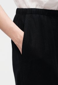 Άτομο που φοράει μαύρο παντελόνι με ελαστική μέση, με το χέρι να ακουμπάει στην πλαϊνή τσέπη, μπεζ τοπ μέσα από το παντελόνι.