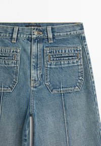 Blue denim jeans feature a high waist, front button closure, and two back pockets with horizontal stitching. The fabric has a worn look.