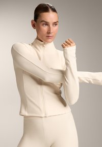 Light beige zip-up jacket made from ribbed material, featuring long sleeves with extended cuffs, ideal for active wear.