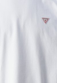 Weißes Baumwoll-T-Shirt mit rundem Halsausschnitt. Verfügt über ein kleines rotes und schwarzes dreieckiges Logo-Patch auf der linken Brust. Glatte Textur.