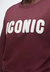 Sweat-shirt marron avec de grandes lettres blanches texturées formant "ICONIC" sur la poitrine.