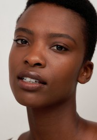 Primer plano de un rostro con piel oscura y lisa, cabello corto y texturizado, maquillaje natural que resalta los labios y los ojos, contra un fondo claro.
