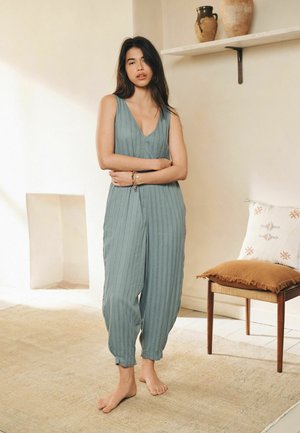 Tânără stă desculță într-un salopetă albastră fără mâneci, într-o cameră minimală bej cu un scaun, o pernă și ceramică pe raft.