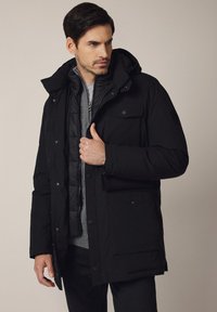 Czarna kurtka puchowa z kapturem, z zamkiem błyskawicznym i zapięciem na napy, przednimi kieszeniami i gładką teksturą, nałożona na szary sweter.