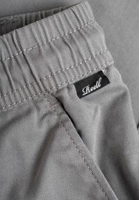 Pantalones cortos de tela gris con una cintura cosida, con una etiqueta negra que dice "Reell". Tela texturizada y costuras visibles.