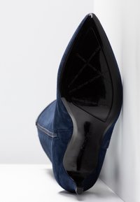 Botte à talon haut en daim bleu marine à bout pointu avec semelle noire embossée du logo "Calvin Klein", photographiée du côté de la semelle.