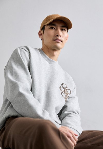 Felpa grigia con un design floreale ricamato marrone sul lato sinistro, abbinata a pantaloni marroni e a un cappellino beige. Design semplice e informale.