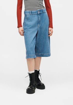 Person som har på seg løse denimshorts til knærne, svarte platå-støvler med lisser og hvite sokker, en rød genser med langermet, og en svart- og hvitstripet skjorte.