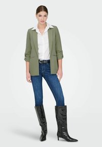 Blazer vert olive sur mesure, chemise blanche à col, jean bleu et bottes noir verni montant jusqu'aux genoux. Les bottes ont un bout pointu et une surface texturée.