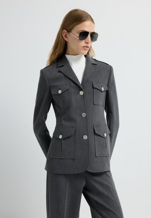 Grijze op maat gemaakte blazer met vier opgestikte zakken, zilveren knopen en een gestructureerde kraag, gecombineerd met grijze broek en een witte coltrui.