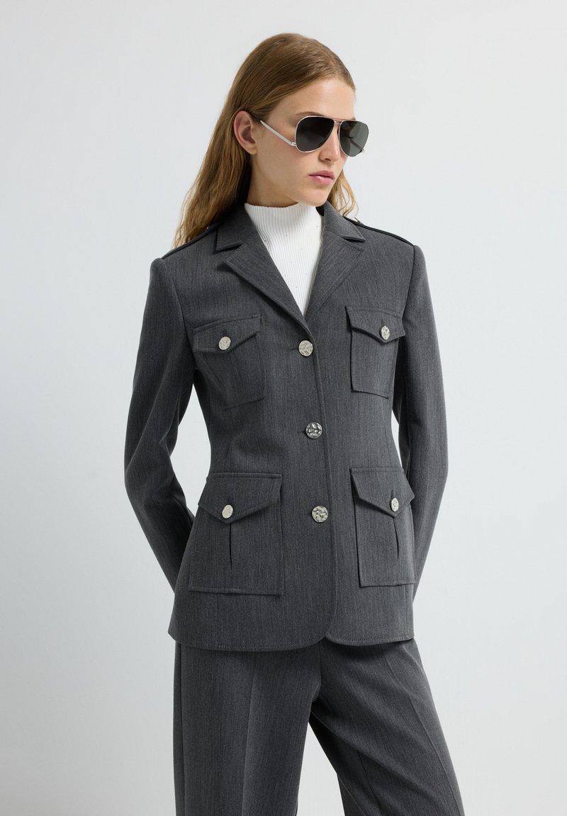 Blazer gris a medida con cuatro bolsillos de parche, botones plateados y un cuello estructurado, combinado con pantalones gris y un suéter de cuello alto blanco.