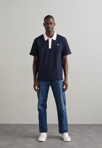 Marineblå polo skjorte med hvit krage og logo, båret med blå jeans og hvite sneakers. Korte ermer, klassisk passform. Enkelt design.