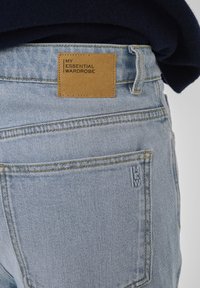 Světlé modré džíny z denimu, pohled zezadu, s hnědou koženou nášivkou označenou "Můj základní šatník" a malou značkovou štítkem na kapse.