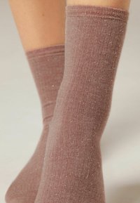 Gros plan sur des jambes portant des chaussettes côtelées rosebrun mi-mollet avec du peluchage visible et une texture de tissu doux.