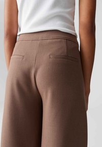 Bruine op maat gemaakte broek met een gladde textuur, voorzien van twee achterzakken en een gestructureerde tailleband, gecombineerd met een witte geribbelde top.