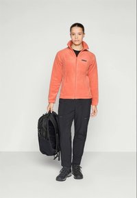 Veste en polaire orange avec un col montant, fermeture éclair noire et logo, associée à un pantalon noir et des bottes robustes. Sac à dos noir tenu à la main gauche.
