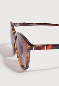 Lunettes de soleil écaille de tortue avec une finition brillante, montures surdimensionnées et incurvées, verres dégradés marron, et détails en métal doré sur les branches.