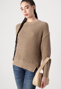 Beige knit sweater with a textured pattern, ribbed hem, and side slits, paired with a small beige handbag and blue jeans.