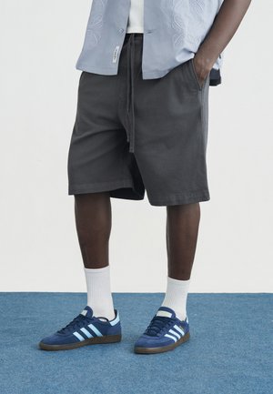 Persona con pantaloncini neri, calzini bianchi e scarpe Adidas blu navy in piedi su un pavimento coperto da moquette blu.