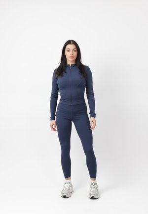 Frau, die nach vorne schaut, trägt eine marineblaue, eng anliegende, mit Reißverschluss versehene Jacke und Leggings sowie weiße Turnschuhe vor weißem Hintergrund.