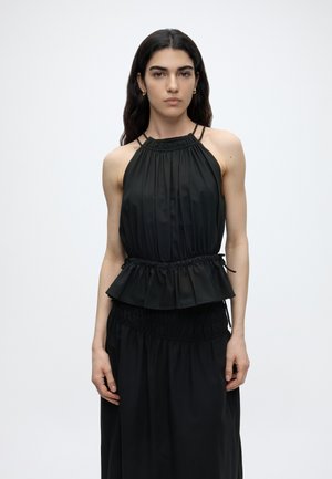 Frau mit langen dunklen Haaren, die ein ärmelloses schwarzes Neckholder-Top mit geraffter Taille und einen passenden schwarzen Rock trägt, stehend vor einem schlichten Hintergrund.